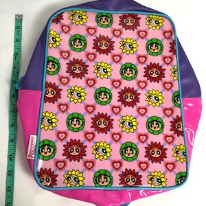 May include: A pink, purple, and blue backpack with a Powerpuff Girls design. The backpack features a repeating pattern of the Powerpuff Girls' faces in sunflowers with hearts on a pink background.