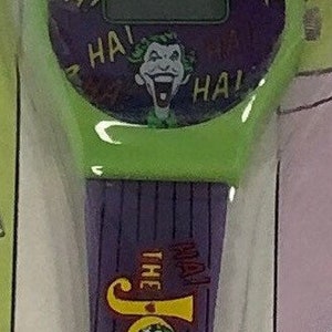 K&ouml;nnte beinhalten: Eine gr&uuml;n-violette Armbanduhr mit digitaler Anzeige und einem Joker-Motiv. Das Zifferblatt der Uhr hat die Worte "HA!" am Rand gedruckt. Das Armband hat die Worte "The Joker" aufgedruckt.