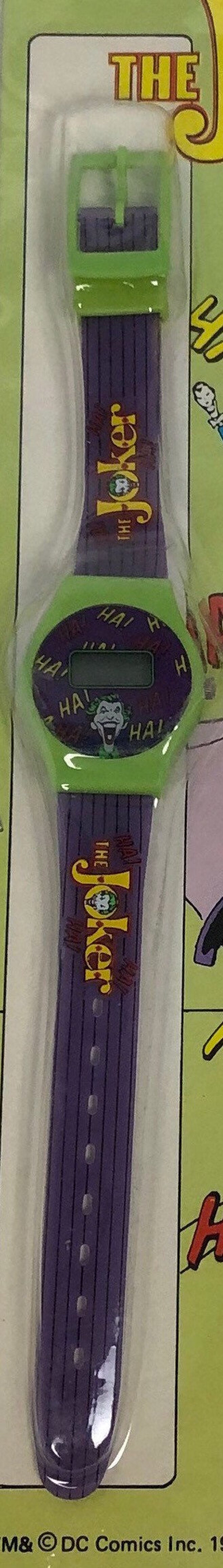 K&ouml;nnte beinhalten: Eine gr&uuml;n-violette Armbanduhr mit digitaler Anzeige und einem Joker-Motiv. Das Zifferblatt der Uhr hat die Worte "HA!" am Rand gedruckt. Das Armband hat die Worte "The Joker" aufgedruckt.