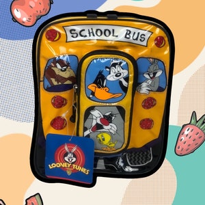 May include: A yellow school bus backpack with a clear plastic front featuring characters from Looney Tunes. The characters include Bugs Bunny, Daffy Duck, Tweety Bird, Sylvester the Cat, and Tasmanian Devil. The backpack has a black zipper and a blue tag with the Looney Tunes logo.