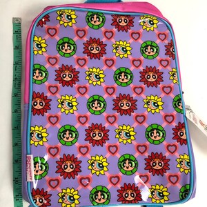 May include: A purple and pink backpack with a repeating pattern of sunflowers and hearts. The sunflowers have the faces of the Powerpuff Girls. The backpack has a blue zipper and a blue strap.