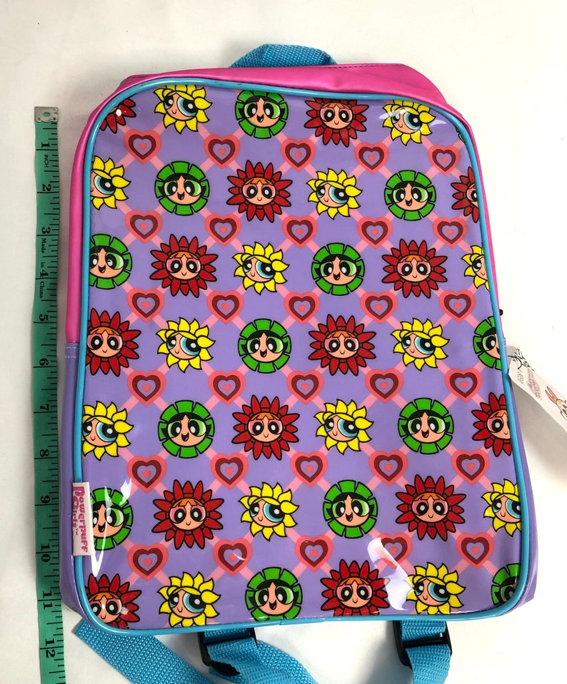 May include: A purple and pink backpack with a repeating pattern of sunflowers and hearts. The sunflowers have the faces of the Powerpuff Girls. The backpack has a blue zipper and a blue strap.