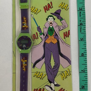 K&ouml;nnte beinhalten: Eine gr&uuml;n-violette Digitaluhr mit einem Joker-Charakterdesign. Das Zifferblatt der Uhr hat ein digitales Display und das Armband tr&auml;gt den Schriftzug "The Joker". Die Verpackung ist gr&uuml;n und zeigt ein Comicbild des Jokers mit den Worten "The Joker Digital Watch" und "Ha!" darauf. TM&&copy; DC Comics Inc. 1989.