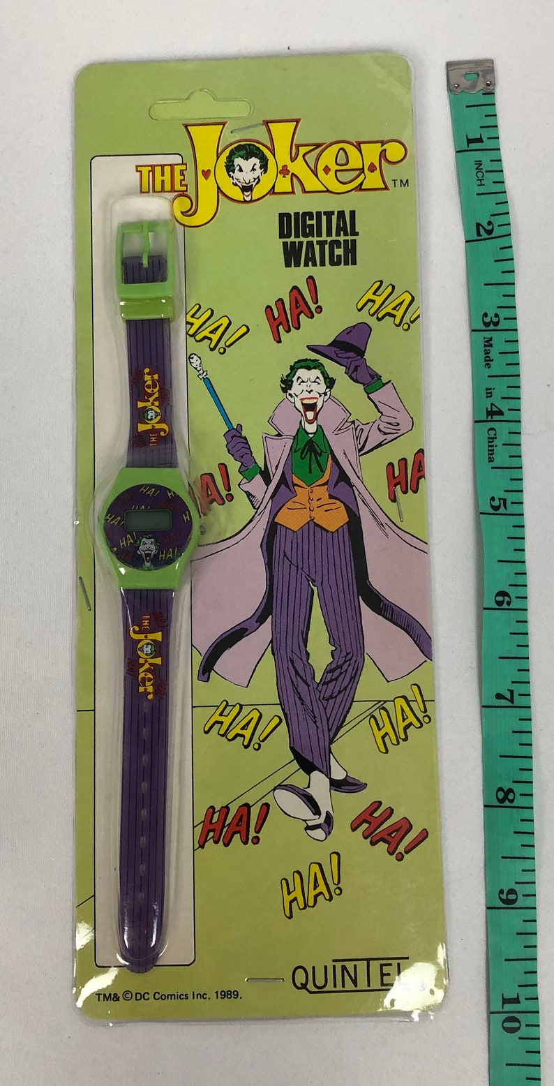 K&ouml;nnte beinhalten: Eine gr&uuml;n-violette Digitaluhr mit einem Joker-Charakterdesign. Das Zifferblatt der Uhr hat ein digitales Display und das Armband tr&auml;gt den Schriftzug "The Joker". Die Verpackung ist gr&uuml;n und zeigt ein Comicbild des Jokers mit den Worten "The Joker Digital Watch" und "Ha!" darauf. TM&&copy; DC Comics Inc. 1989.