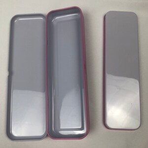 May include: Three empty rectangular tin boxes with a pink trim. The boxes are open and have a silver interior.
