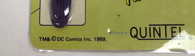 K&ouml;nnte beinhalten: Ein lila-wei&szlig;es Spielzeugtelefon aus Plastik mit gelbem Hintergrund. Das Telefon hat ein schwarzes Zifferblatt und ein schwarzes Kabel. Die Verpackung enth&auml;lt den Text "TM & &copy; DC Comics Inc. 1989." und "QUINTEL".