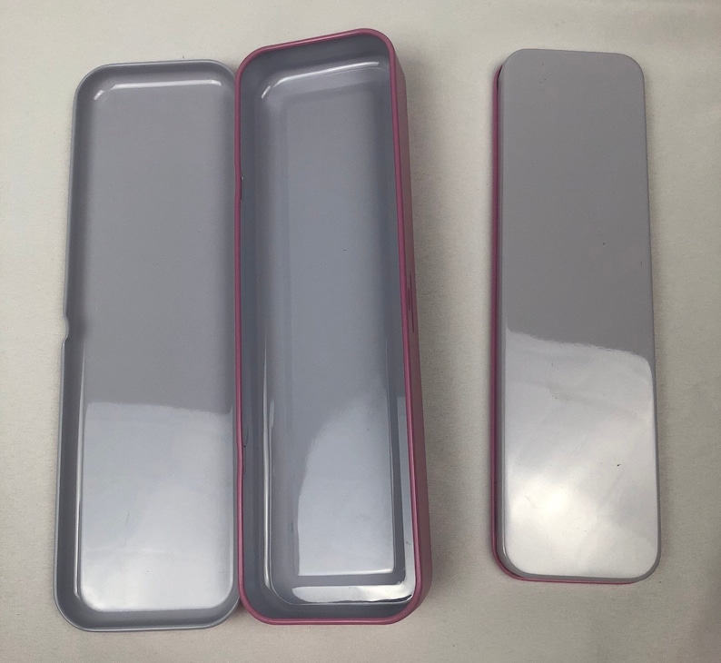 May include: Three empty rectangular tin boxes with a pink trim. The boxes are open and have a silver interior.