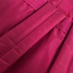 May include: Close-up of a deep red backpack strap with a woven design. The strap is made of a smooth, nylon-like material.