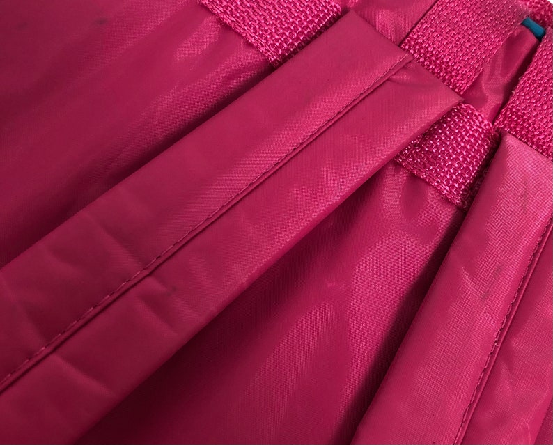May include: Close-up of a deep red backpack strap with a woven design. The strap is made of a smooth, nylon-like material.
