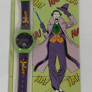 K&ouml;nnte beinhalten: Eine gr&uuml;n-violette Digitaluhr mit einem Bild des Jokers auf dem Zifferblatt. Die Uhr ist in einem gr&uuml;n-gelben Karton verpackt, auf dem ein Bild des Jokers und die Worte "The Joker Digital Watch" zu sehen sind.