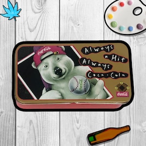 Puede incluir: Una fiambrera vintage de Coca-Cola de metal con un oso polar que lleva una gorra de béisbol y sostiene una pelota de béisbol. La fiambrera es dorada con detalles rojos y tiene el texto "Always a Hit Always Coca-Cola" en la parte delantera.