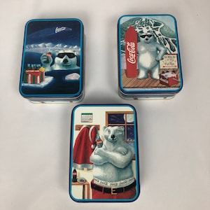 May include: Three vintage Coca-Cola collectible tins featuring a polar bear. One tin shows the bear holding a Coca-Cola bottle, another shows the bear on a snowboard, and the third shows the bear wearing a Santa hat and a Coca-Cola belt.