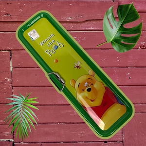 May include: A green and yellow pencil case with a Winnie the Pooh design. The case features a picture of Winnie the Pooh with a bee on his nose, a bee flying, and a sun.