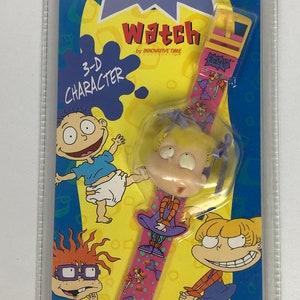 May include: A pink and yellow Nickelodeon Rugrats watch with a 3-D character of Angelica Pickles. The watch features a pink band with a polka dot pattern and a white background. The watch face is not visible. The packaging includes a warning about choking hazards for children under 3 years old.