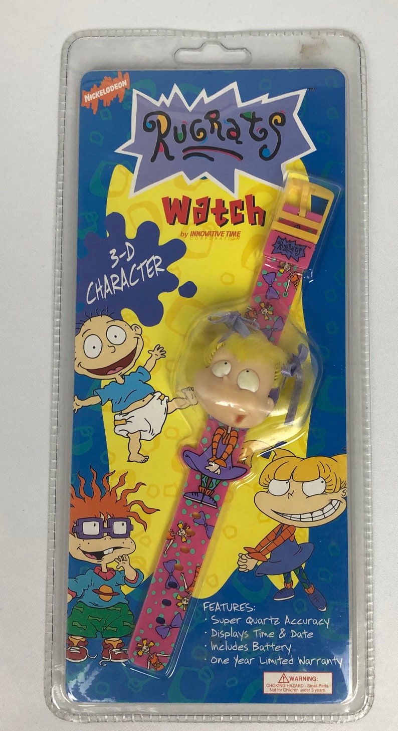 May include: A pink and yellow Nickelodeon Rugrats watch with a 3-D character of Angelica Pickles. The watch features a pink band with a polka dot pattern and a white background. The watch face is not visible. The packaging includes a warning about choking hazards for children under 3 years old.