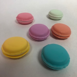 Könnte beinhalten: Sechs pastellfarbene macaronförmige Behälter mit weißem Frosting. Die Farben sind rosa, lila, orange, gelb, blau und grün.