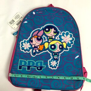 May include: A pink and blue backpack with a Powerpuff Girls design. The backpack features the three girls, Blossom, Bubbles, and Buttercup, with the words "PPG" printed on the front. The backpack is made of a shiny, vinyl-like material.