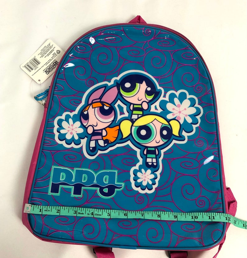 May include: A pink and blue backpack with a Powerpuff Girls design. The backpack features the three girls, Blossom, Bubbles, and Buttercup, with the words "PPG" printed on the front. The backpack is made of a shiny, vinyl-like material.