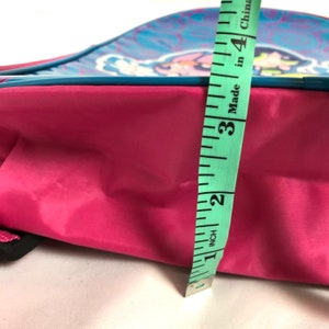 May include: A pink backpack with a blue and white design featuring cartoon characters. The backpack is being measured with a tape measure that reads "Made in China" and shows a measurement of 3 inches.