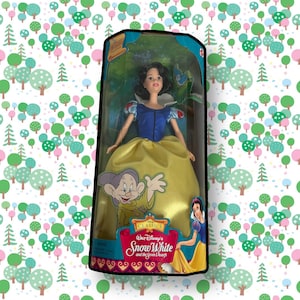 Op de afbeelding: Een vintage Disney Classics pop van Sneeuwwitje in haar iconische gele en blauwe jurk. De pop is verpakt in een doorzichtige plastic doos met een kleurrijke illustratie van Sneeuwwitje en de Zeven Dwergen. De doos bevat ook de tekst "Disney Classics Sneeuwwitje en de Zeven Dwergen".