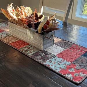 May include: A wooden rectangular box filled with dried flowers and pinecones sits on a patchwork table runner. The runner features squares of various patterns in red, brown, and teal. The table is dark wood.