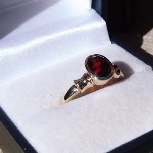 Peut inclure: Une bague en or avec une grosse pierre précieuse ovale rouge foncé, présentée dans un écrin à bijoux bleu avec un intérieur en satin blanc. La bague a des éléments décoratifs.