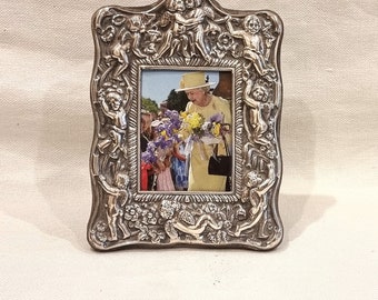 May include: A silver picture frame with an ornate design featuring cherubs and floral motifs. The frame holds a photo of a woman in a yellow dress and hat, holding flowers.
