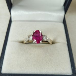 May include: A gold ring featuring a large oval ruby and two smaller white stones. The ring is set in a gold band and presented in a blue and white ring box. The ruby is the focal point.