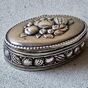 May include: An antique silver-toned oval box with a detailed floral design on the lid. The box features a beaded border and ornate patterns around the sides. A vintage jewellery or trinket box.