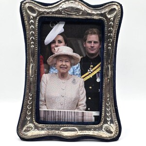 May include: A silver picture frame with a blue velvet backing. The frame features a photo of Queen Elizabeth II, Prince Harry, and Catherine, Duchess of Cambridge.