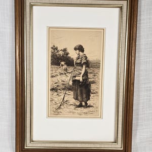 May include: Framed sepia-toned print of a woman in a field, holding a farming tool. The artwork is set in a brown wooden frame with a white mat and a linen-textured inner border. The print depicts a rural scene.