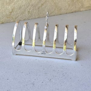 Peut inclure: Un porte-toast argenté avec six fentes et une poignée. Le support a une base rectangulaire et des séparateurs arqués. La poignée est une petite boucle. Le support est en métal brillant.