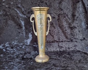 May include: A tall, antique brass vase with two curved handles and decorative floral and leaf motifs. The vase has a wide, flared top and a round base. The brass has a warm, golden hue, and the design is Art Nouveau in style.