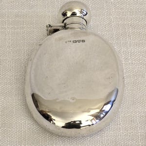 May include: A round, polished silver flask with a smooth, reflective surface and a rounded screw-top cap. The body of the flask features a series of small, stamped hallmarks near the top, indicating its metallic composition and origin.