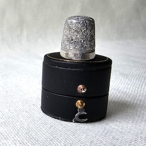 May include: A silver thimble with a floral design is displayed on a black, round, two-part box. The thimble is intricately patterned and has text markings. The box features gold trim and a clasp for closure.