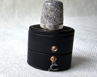 May include: A silver thimble with a floral design is displayed on a black, round, two-part box. The thimble is intricately patterned and has text markings. The box features gold trim and a clasp for closure.