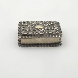 May include: A small, rectangular silver box. The lid features detailed floral and scrollwork, with a raised central design and a beaded border. The sides are plain silver. The box is likely for jewellery or small keepsakes.