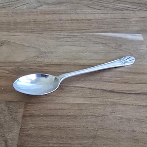 May include: A silver-toned metal spoon with a smooth, oval-shaped bowl and a slender handle. The end of the handle features a decorative, fan-shaped engraving consisting of several radiating lines that create a distinct, textured pattern.