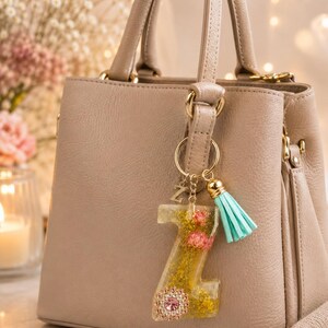 Puede incluir: Un bolso de cuero beige con un llavero de resina en forma de letra Z. El llavero contiene flores secas rosas y amarillas, purpurina dorada y una pequeña flor de pedrería rosa, acentuado con una borla de color turquesa y un dije de letra Z.