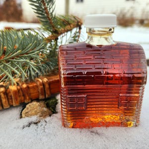May include: A small, house-shaped glass bottle filled with amber liquid, likely syrup, with a white cap. The bottle sits in snow, with a pine branch and wooden element in the background. The bottle has a brick-like texture.