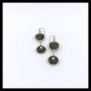 May include: A pair of dangle earrings with a gold-colored hook and chain. Each earring features a dark brown, heart-shaped stone above a circular stone with a silver-colored bead in the center. The earrings are displayed on a white background.
