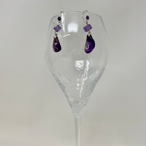 May include: A pair of amethyst earrings with silver-colored hooks. Each earring features a small, square amethyst bead, a teardrop-shaped amethyst, and a small round amethyst bead. The earrings are displayed in a clear wine glass against a white background.