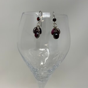 May include: A pair of silver earrings with a cluster of dark red, oval-shaped gemstones. The earrings are suspended from a silver hook and are displayed in front of a clear wine glass.