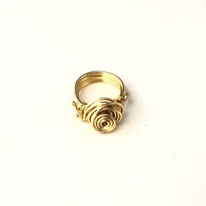 May include: A gold-colored wire ring featuring a rose design. The ring's band has multiple parallel wires, and the rose is crafted with spiraling wire. The ring is set against a white background.