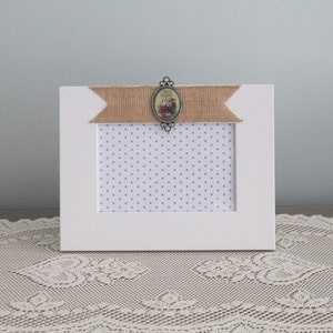 May include: White picture frame with a burlap ribbon and a decorative oval cameo with a floral design. The frame has a white background with a gray polka dot pattern.
