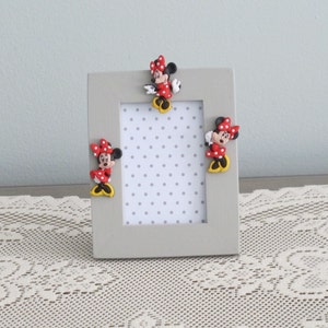 May include: A gray picture frame with a white background and three red and white polka dot Minnie Mouse figures attached to the frame.