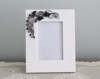 May include: White picture frame with a silver and black floral design on the top left corner. The frame has a white mat and a clear glass front.