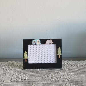 May include: Black picture frame with a white background and a polka dot pattern. The frame has a small blue camper, a white tent, and two green trees on the top.