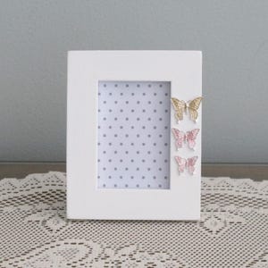 May include: A white picture frame with three decorative butterflies in gold and pink. The frame has a rectangular opening with a polka dot background. The butterflies are arranged vertically on the right side of the frame.