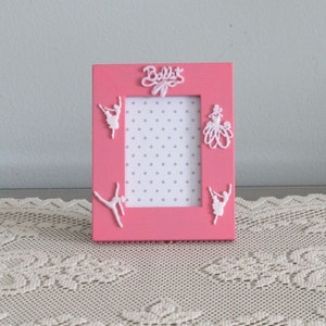 May include: Pink picture frame with white ballerina silhouettes and the word "Ballet" in white letters. The frame has a white polka dot background.